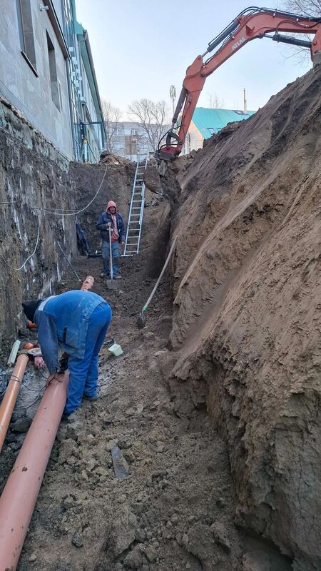 Mélyre ásott közműárok épület alapfala mellett, alján fektetett narancssárga csatornacső, a felszínen forgókotró
