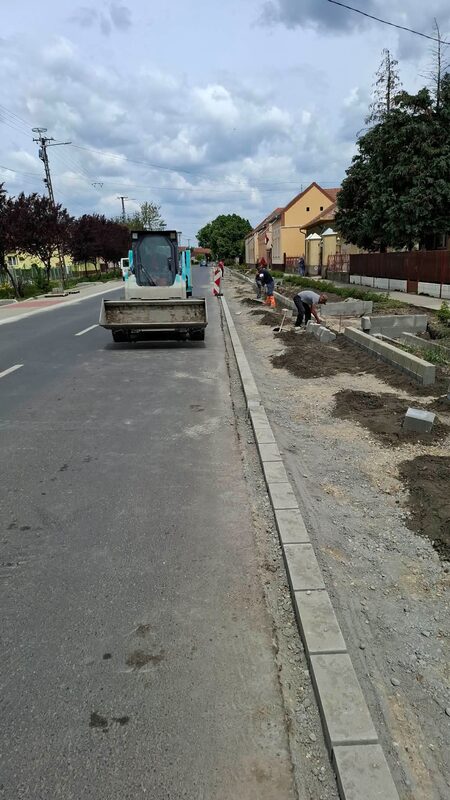Hosszan lefektetett betonszegély az út mentén, mögötte a járdatükör földmunkáján dolgozik a csapat, az úton kompakt rakodógép
